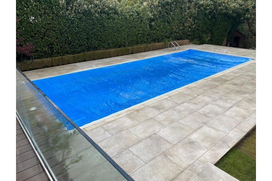 View down from glass-railed balcony to long, blue-lined swimming pool with Frosty Grey porcelain paving surround, by S & D Paving.