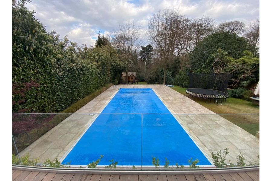 In mature garden, Frosty Grey Porcelain outdoor tiles, laid by S&D Paving, surround long, rectangular blue-lined swimming pool.