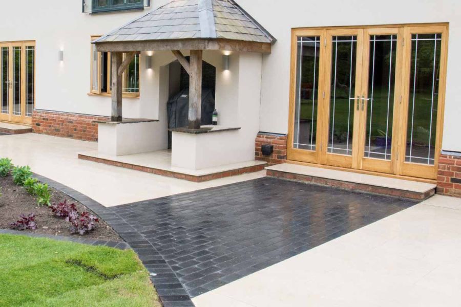 Wooden french doors open onto Midnight Black limestone cobble setts that cross white paving and meet curved edge of lawn and beds.