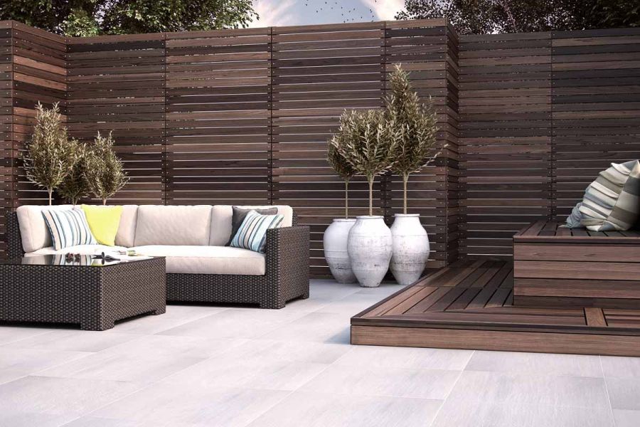 Tall dark wood slatted fence backs matching decked area on Flamed Grey sandstone paving with modular sofa and large pots.