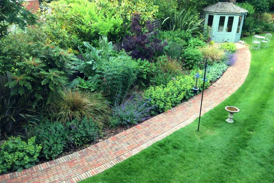 Long, gently curved path separates lawn from deep herbaceous border and leads to summer house with bistro set.