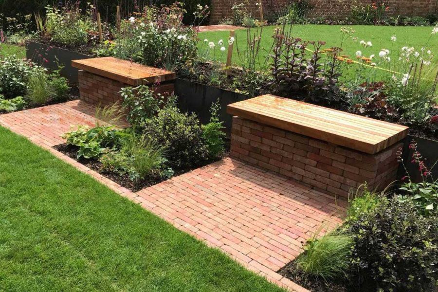 Flower beds on either side of 2 brick-built, wood-topped benches with aprons of Seville clay pavers in front of them.