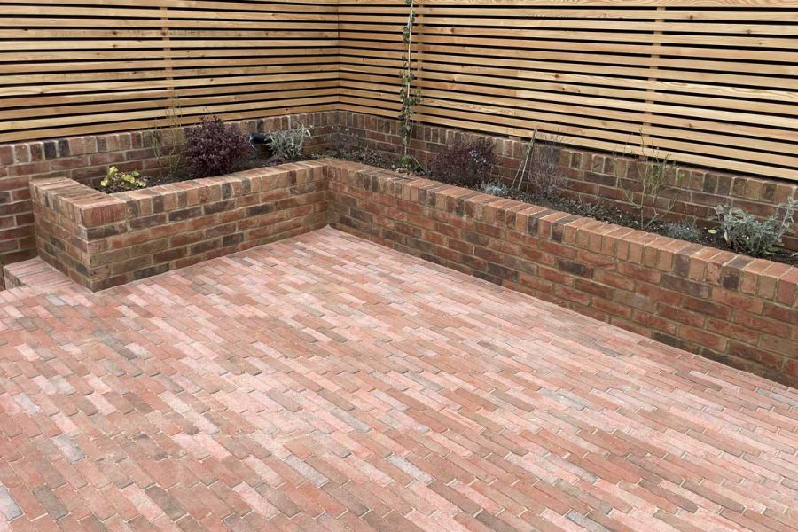 Seville clay paving edged on two sides by brick raised bed in corner of garden with slatted fence on top of low wall.