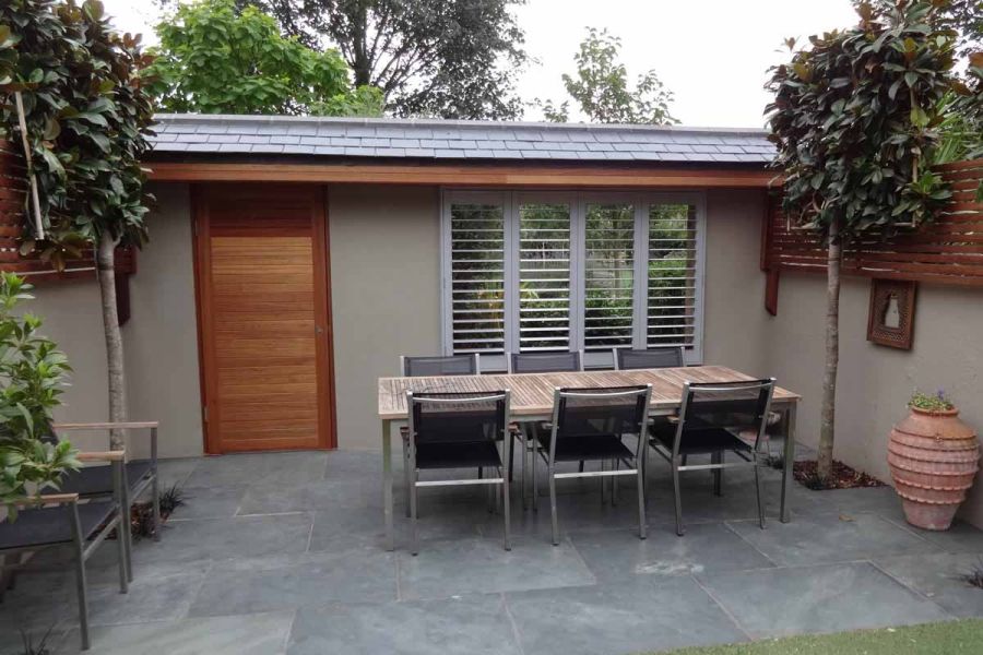 Roofed wall with louvred window and wooden door at back of outdoor dining area paved with Green slate 900x600 slabs.