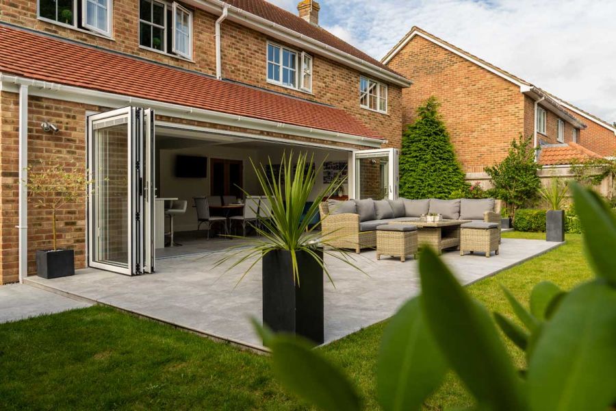 Silver Contro porcelain patio connected to back of house by wide bifold doors, with luxury rattan furniture and square planters.