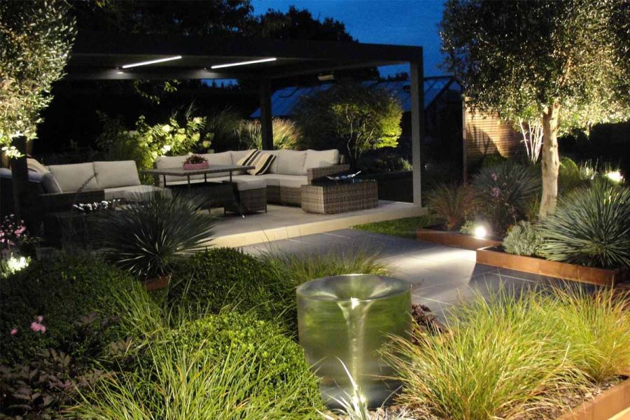 view at night of densely planted back garden with lit up pergola, rattan furniture underneath, all sat on silver contro porcelain paving.