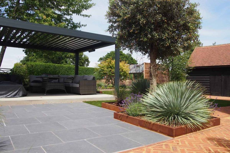 pergola seen in the background of large garden using silver contro porcelain paving. Low level corten planters in the foreground.