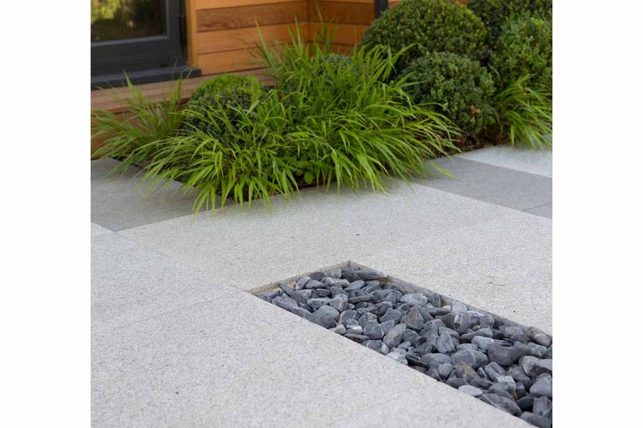 Oblong of grey pebbles set into to Silver Grey granite paving outside wooden building with bed of grasses and topiary balls.