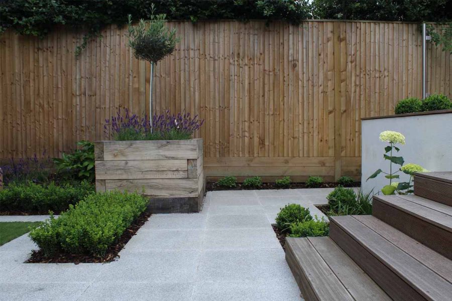 4 wooden steps descend to Silver Grey granite paving with timber sleeper square planter and narrow border against tall fence.
