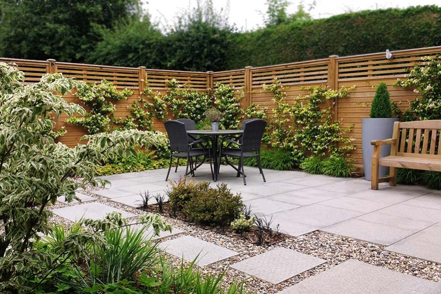 Patio of Silver Grey granite paving in corner of fenced garden, with variegated plants in borders and clipped fir in tall pot.
