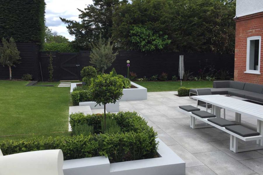 Large Silver grey granite patio with white walled raised beds, hedges and topiary. Lawn on 2 sides. Built by Elite Landscapes.
