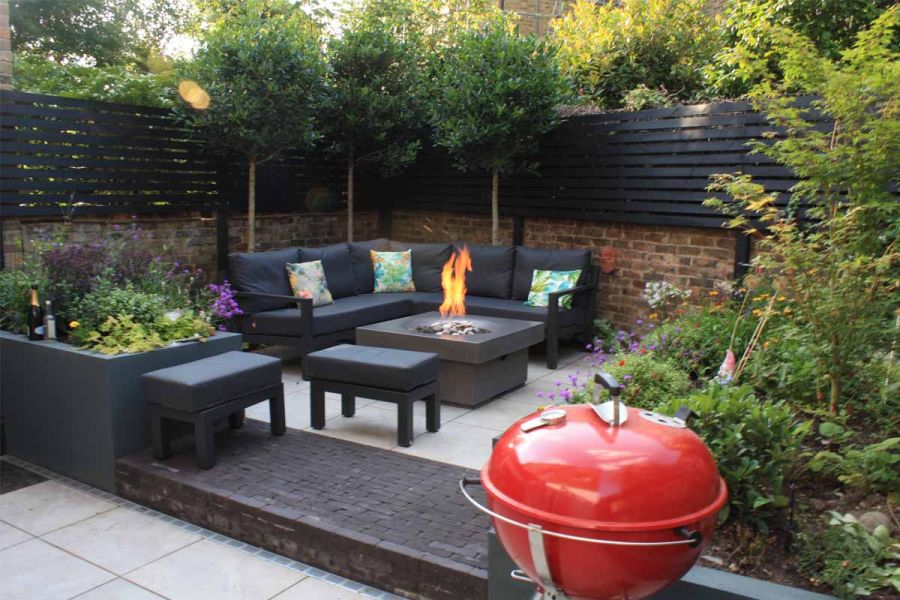 Small walled garden. Wide charcoal clay paver step. Silver-grey porcelain paving with outdoor sofa, fire-table and stools.