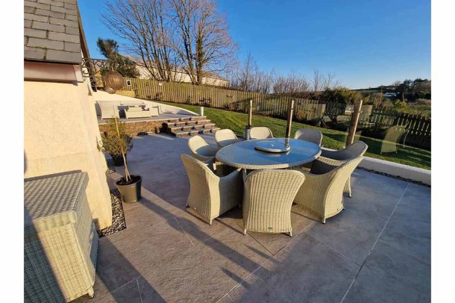 Silver grey porcelain paving shows off texture in large back garden with sloping lawn and rattan furniture.
