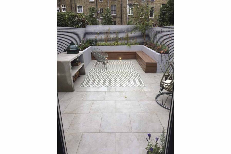 Small back garden with silver-grey porcelain paving, edging rectangle of patterned tiles. Bench attached to raised beds.