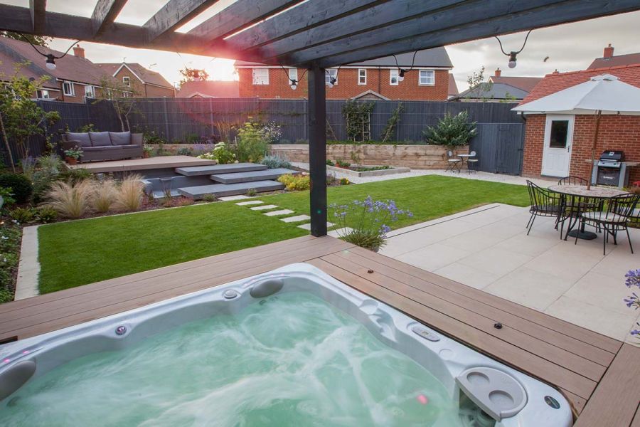 Florence beige porcelain-tiled patio, with hot tub. Paving slabs in lawn lead to raised patio, by Simon Orchard Garden Design.