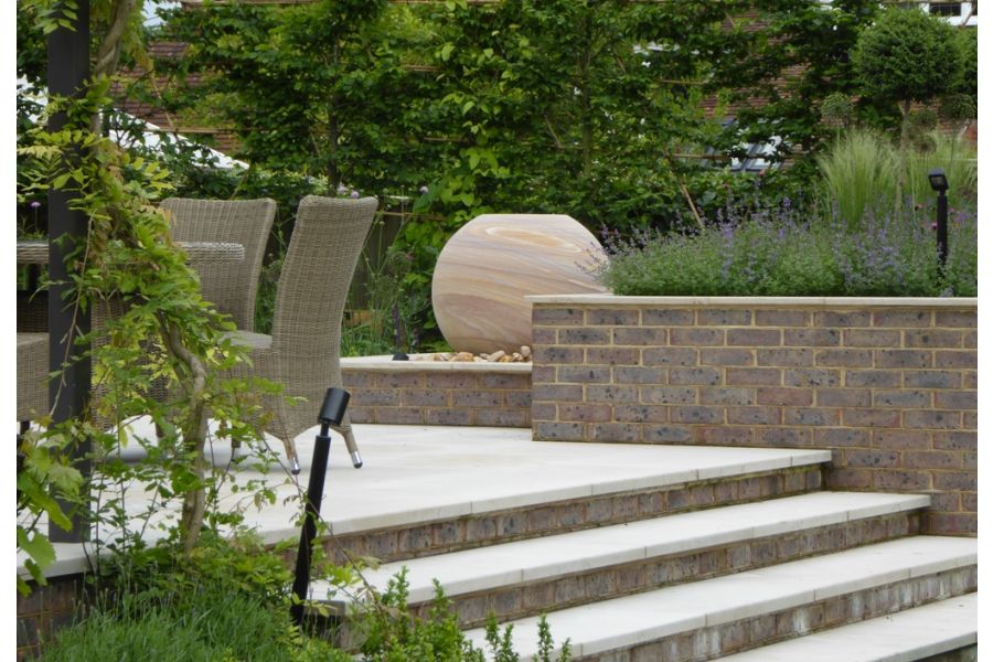 4 Jura Beige sawn limestone steps with brick risers rise to patio with outdoor furniture and spherical water feature on pebbles.