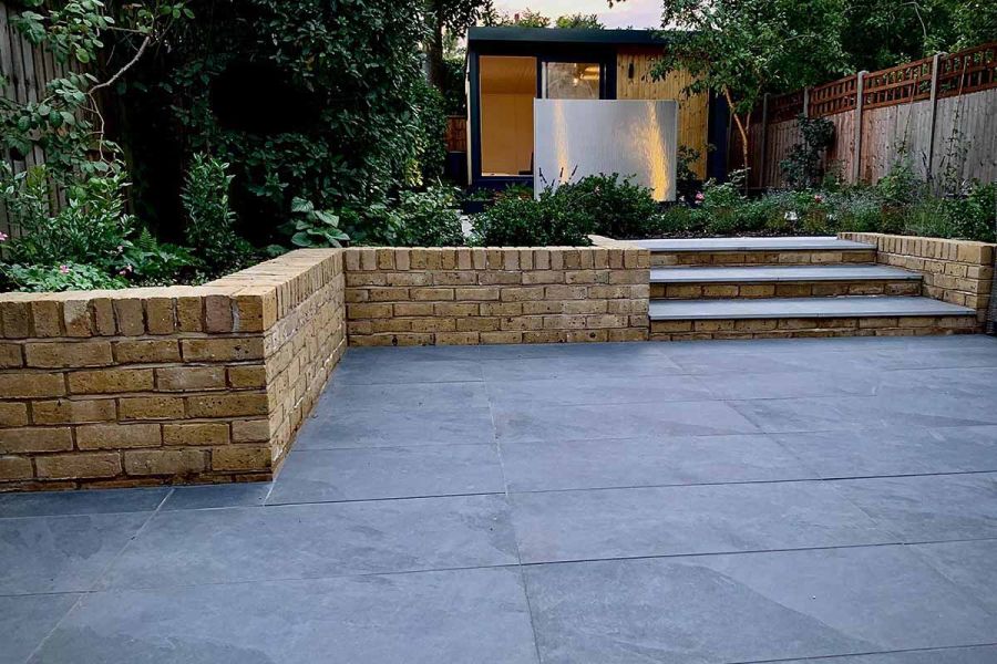 Wide Slab Coke porcelain steps rise between low brick flanking walls from matching paved area to path between planted borders.