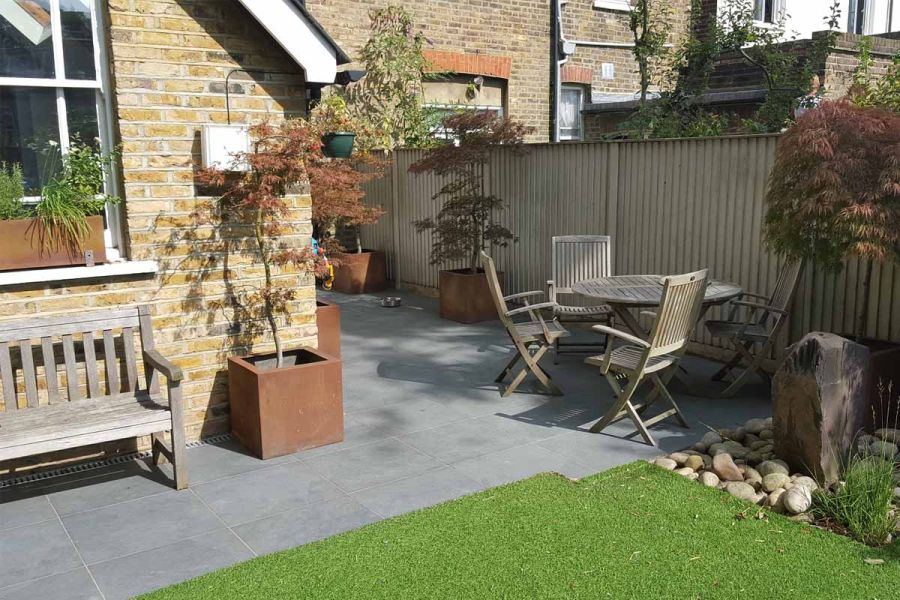 Slab Coke porcelain paving wrapped around corner of house, with trees in corten steel planters. Built by Oakley Landscapes.