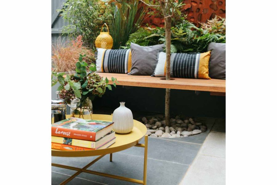 Round yellow coffee table on Slab Coke Porcelain outdoor tiles UK. Behind, small tree grows through hole in wooden bench seat.