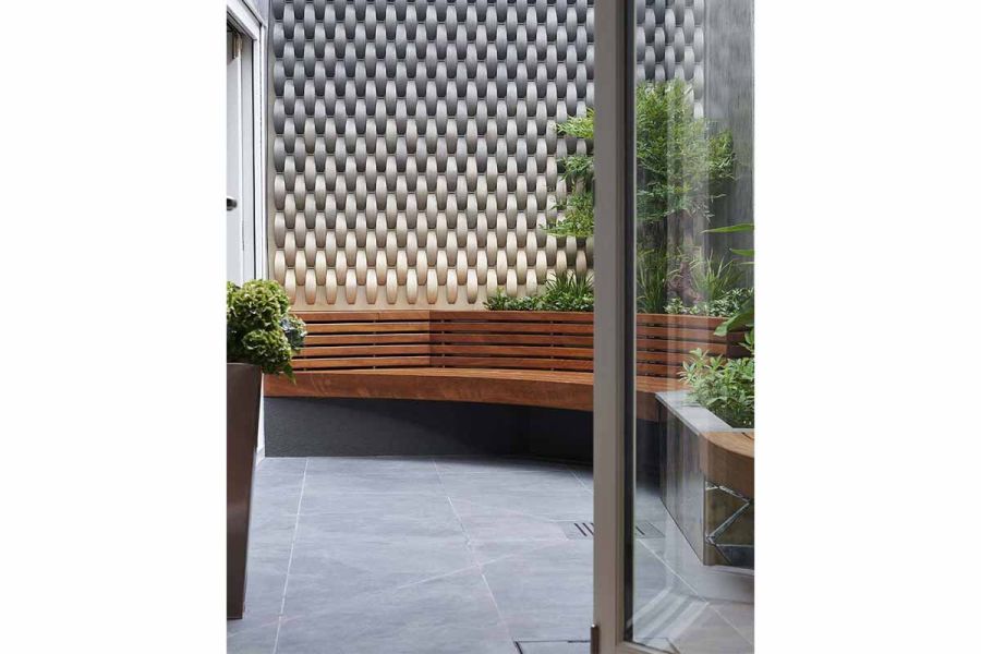 Open door onto small courtyard paved in Slab Coke porcelain, with wooden bench, modern metal screen and planting in raised beds.