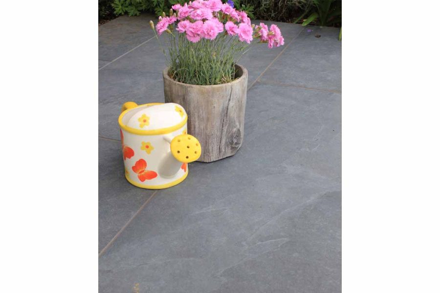 Orange, yellow and cream china watering can sits next to wooden pot of pink carnations on Slab Coke vitrified porcelain paving.