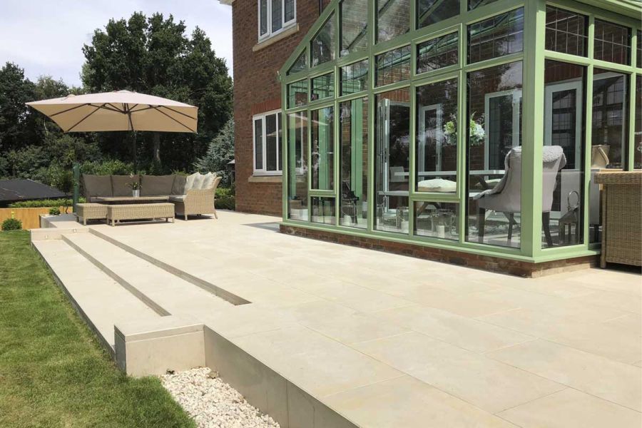 2 wide shallow steps with low flank walls descend to lawn from Slab Khaki porcelain patio with furniture. Built by Aye Gardening.