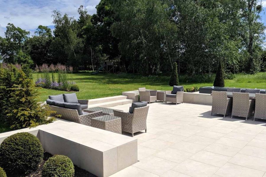 3 sets of luxury rattan furniture on large patio of Slab Khaki porcelain outdoor tiles, with 2 steps up to lawn and mature trees. 