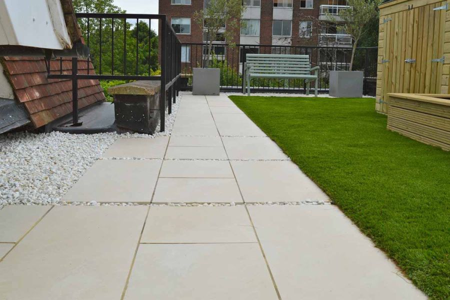Path of Slab Khaki porcelain outdoor tiles leads to wooden bench and planters, between black railings and grassy border. 