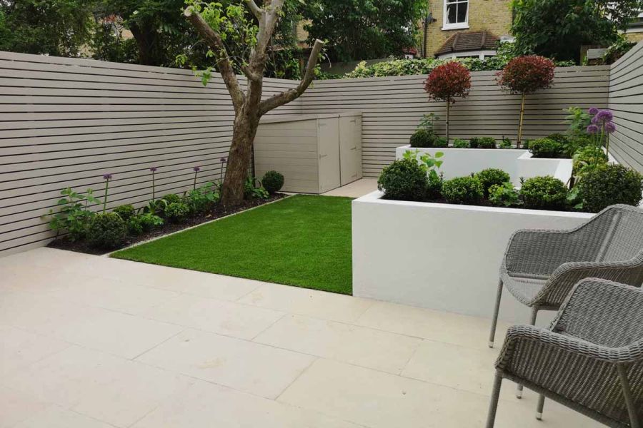 Small back garden with pale slatted fence, white raised beds, lawn and Slab khaki porcelain paved patio. Design by Amelia Bouquet.