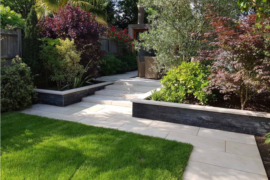 In mature garden, path of Slab Khaki Porcelain Outdoor Tiles UK leads to steps, with flanking wall topped with matching coping.