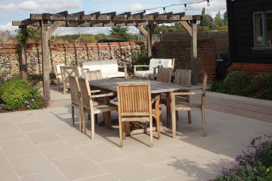 Slab khaki porcelain paving laid next to cobblestone wall has wooden dining table and pergola, with comfy seating area at the back.