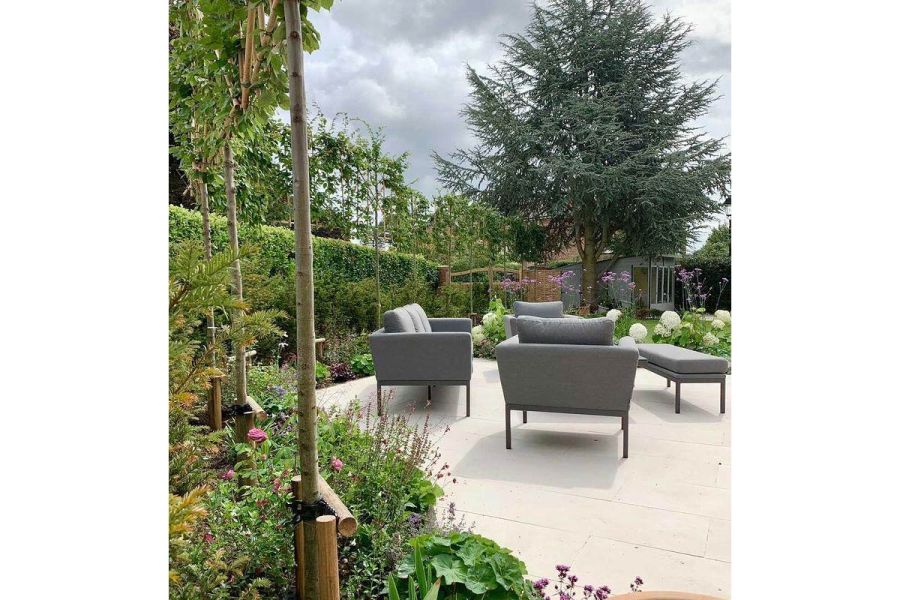 Grey outdoor lounge set on Slab Khaki porcelain paved area edged by verdant borders with tall hedge behind. By Parme Garden Design.