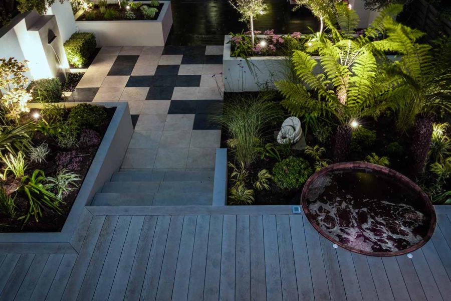 Courtyard with composite decking patio. Slab Khaki porcelain steps descend to two-tone paving with water feature and raised beds.