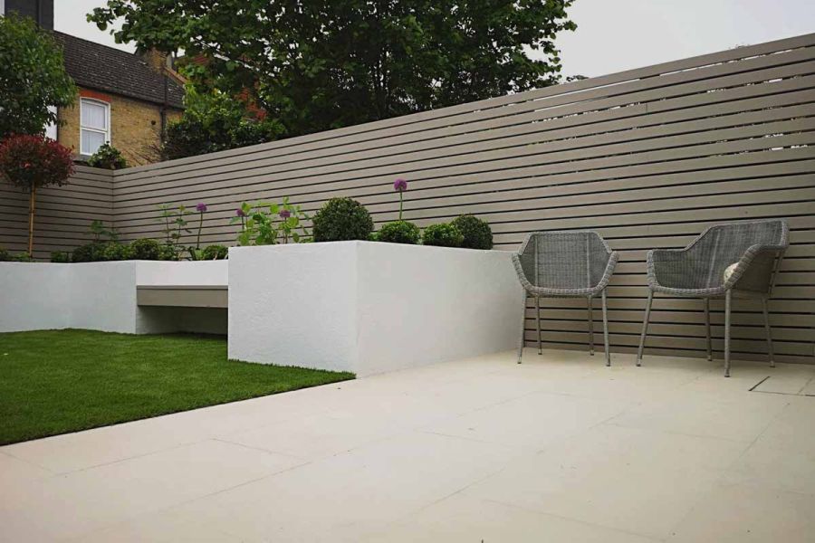 2 rattan chairs sit on Slab Khaki vitrified porcelain tiles next to mushroom-coloured slatted fence and white-rendered raised beds.