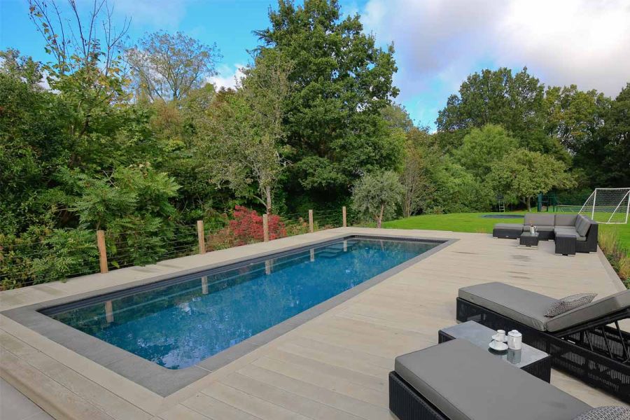 Sun loungers on Smoked Oak Millboard decking surround of swimming pool with stone coping edges, in lawned garden bordered by trees.