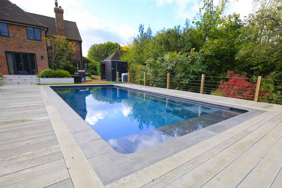 Smoked Oak Millboard decking surrounds rectangular swimming pool with stone coping. Steps in background ascend to house.