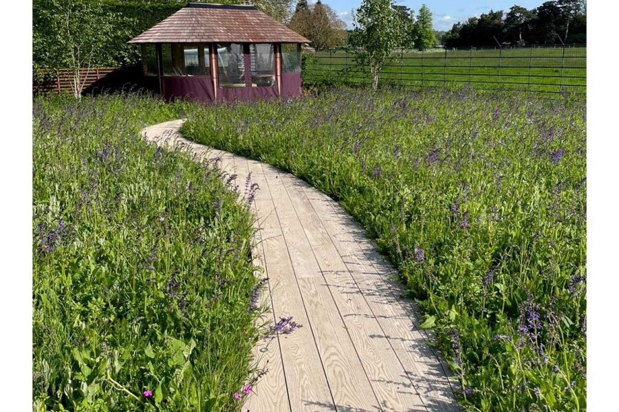 Smoked Oak Millboard decking boards laid longitudinally along sinuous path to tile-roofed hut amidst prairie planting.