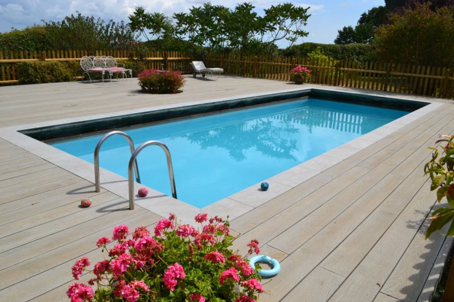 Large fenced patio of Millboard oak smoked composite decking with rectangular swimming pool and pots of pink geraniums.