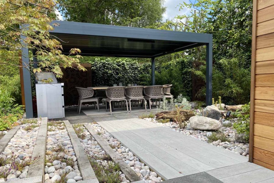 Smoked Oak Millboard composite decking path between beds of large pebbles and dry planting leads to pergola over dining set.