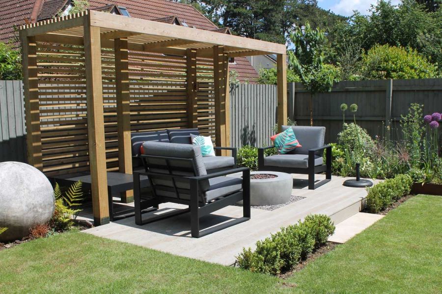 Lawn on 2 sides of small rectangular deck of Millboard Smoked Oak composite boards with chairs, fire pit and wooden pergola.