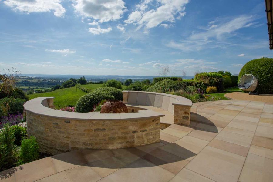 Metal firepit in sunken seating area on edge of wide terrace of buff sawn sandstone with view across English countryside.