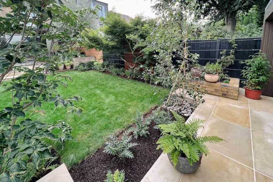 Harvest sawn sandstone slabs laid stack bond as patio with flower bed inset at edge, next to lawn with planted borders.