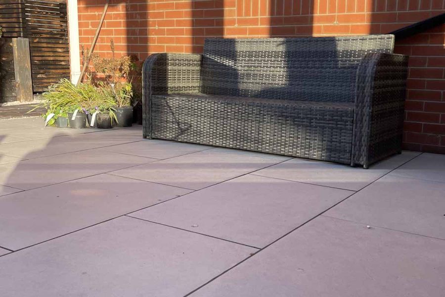 3-seater grey rattan sofa sits on florence grey porcelain paving, sat against side brick wall of house, next to cluster of potted plants.
