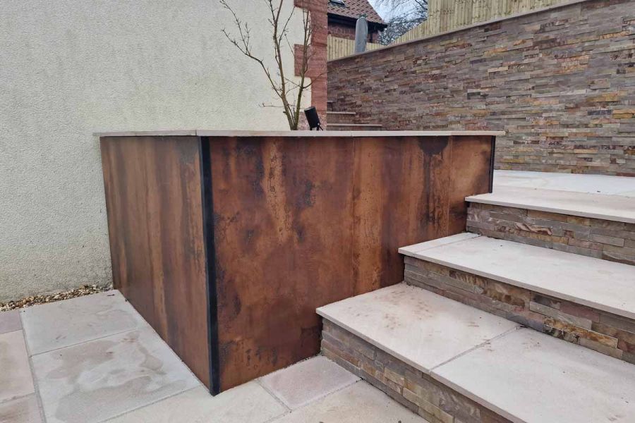 Young tree in deep bed faced with Siena Copper exterior wall cladding. 3 steps rise up one side connecting 2 levels of paving.