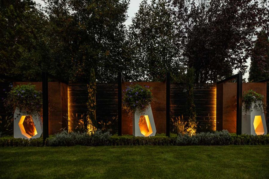 Large asymmetric planters punctured by large hole stand panels of Steel Corten external cladding set at intervals in slatted fence.