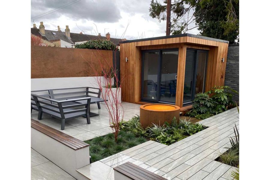 Low white wall topped with panel of Corten Steel external cladding borders paved garden with studio and rusted water feature.