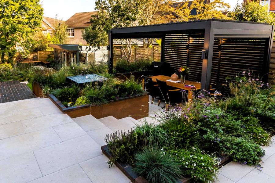 4 steps descend from patio between raised beds, faced with Steel Corten external cladding, to pergola-covered dining area.