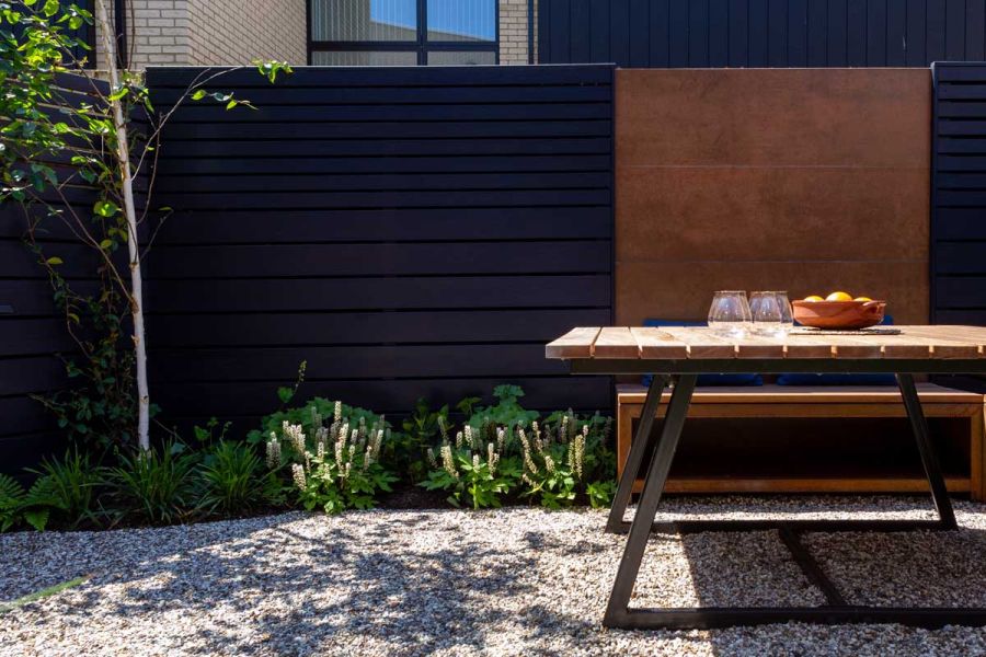 Tall dark fence punctuated by panel of Steel Corten external cladding borders gravel area with silver birch tree, table and bench.