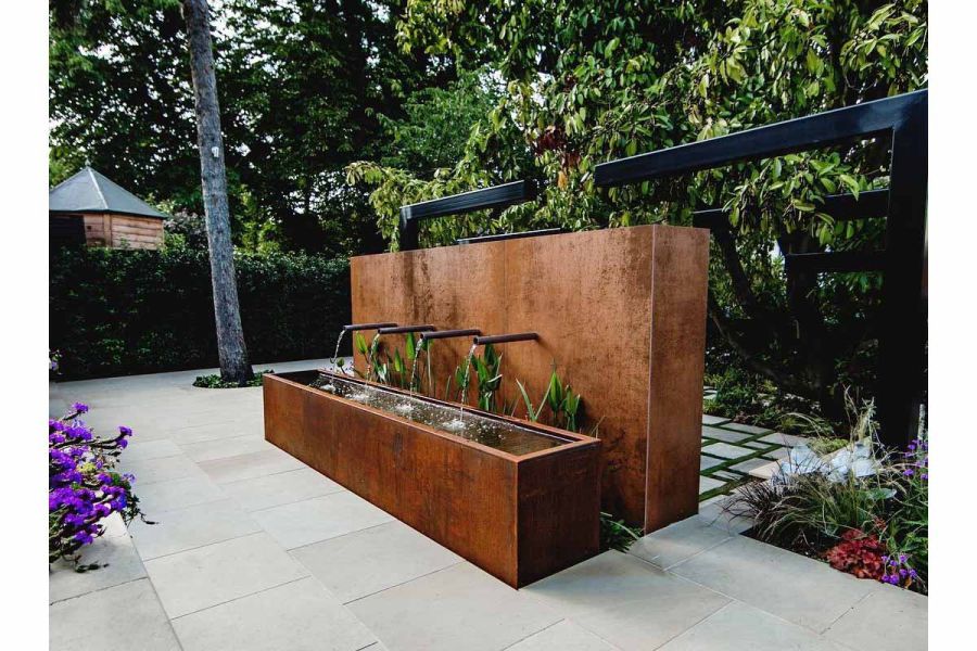 In paved garden, 4 pipes spout water into trough from monolithic water feature, all faced with Steel Corten external cladding.