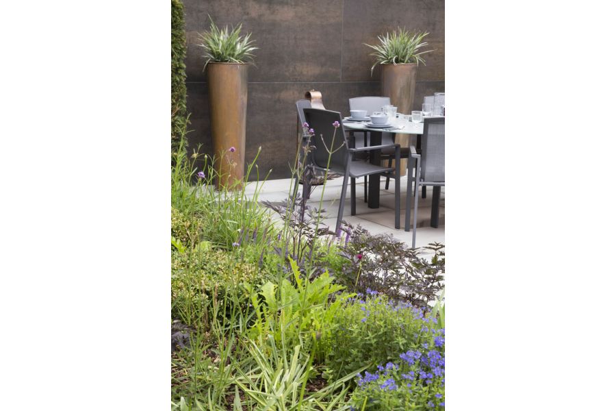 Thickly planted bed next to dining set. 2 tall planted pots stand against wall faced with Steel Dark porcelain cladding panels.
